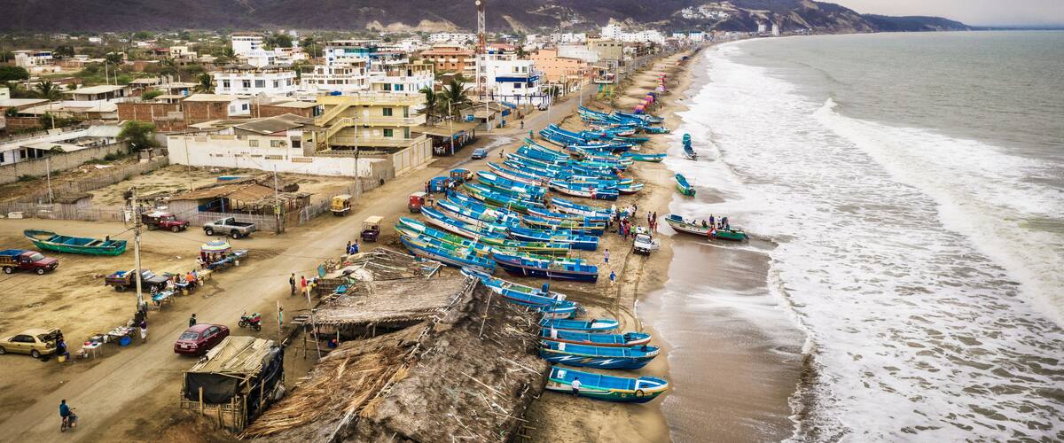 A coastal town with a long row of boats lined up along the beach, a dirt road, and buildings in the background.