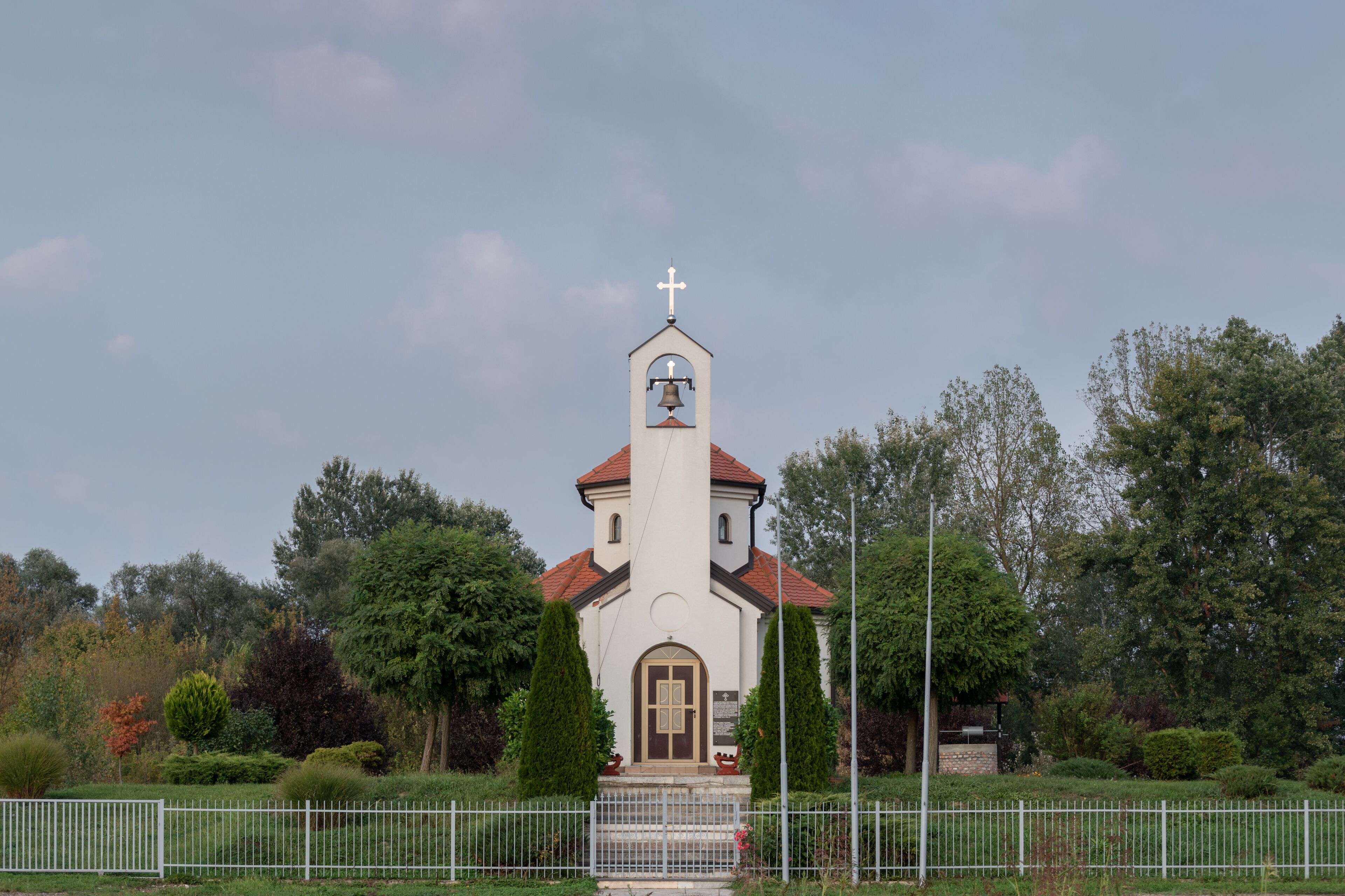 A church with a bell tower and a cross on top, surrounded by trees and a white fence.