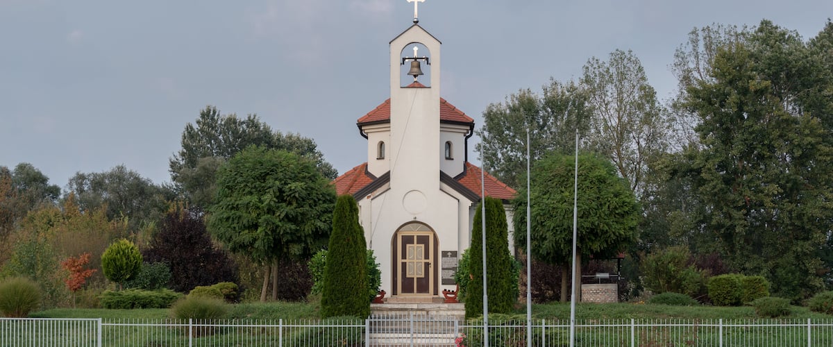 A church with a bell tower and a cross on top, surrounded by trees and a white fence.