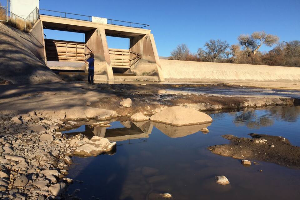 Una presa de concreto con una persona de pie sobre ella, una orilla del río pedregosa y un cielo azul despejado.