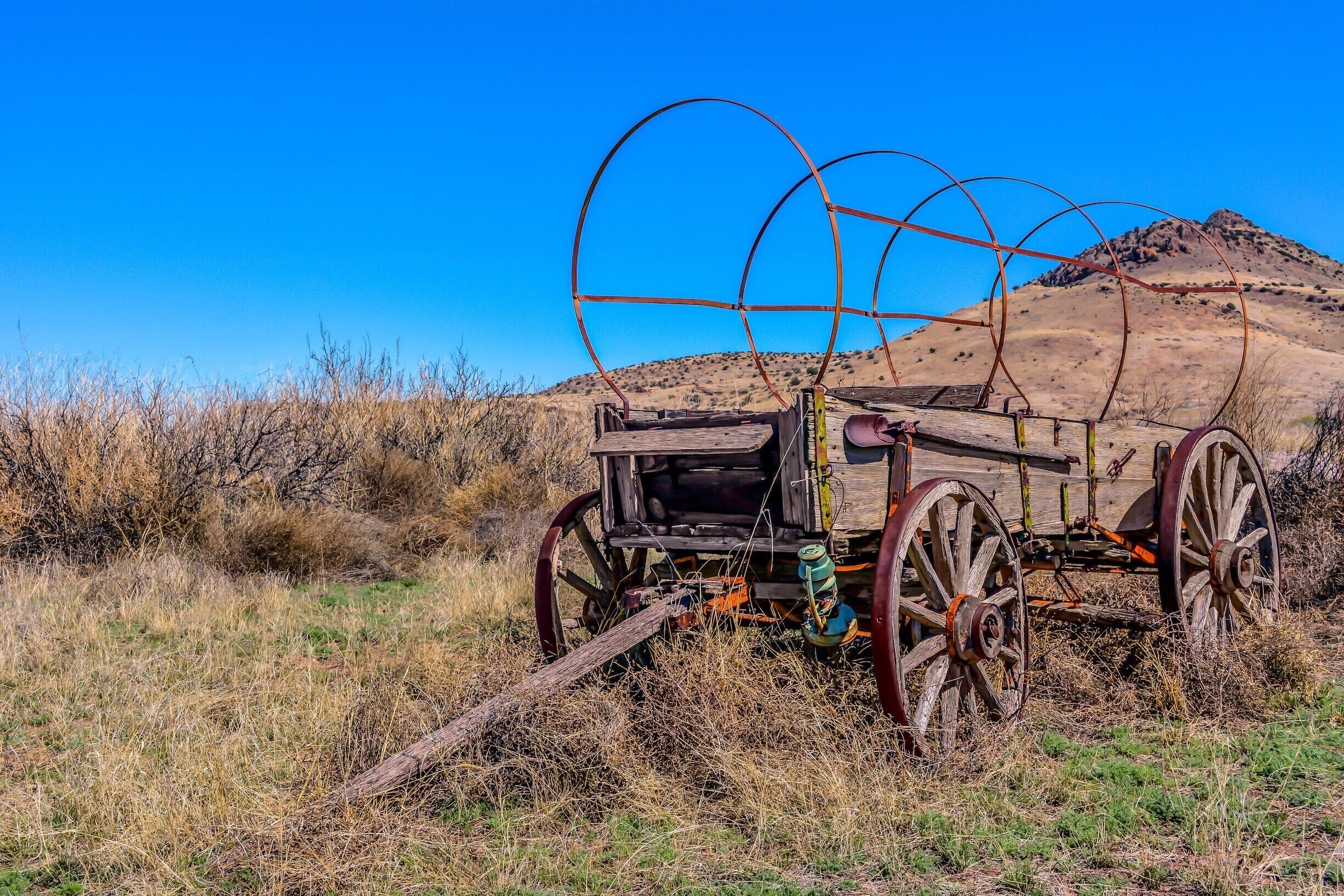 An old, rusted wagon with large wheels in a dry, grassy field with a clear blue sky and a hill in the background.