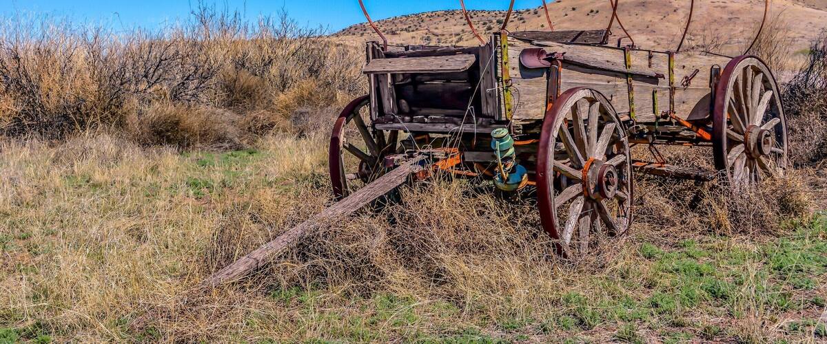 An old, rusted wagon with large wheels in a dry, grassy field with a clear blue sky and a hill in the background.