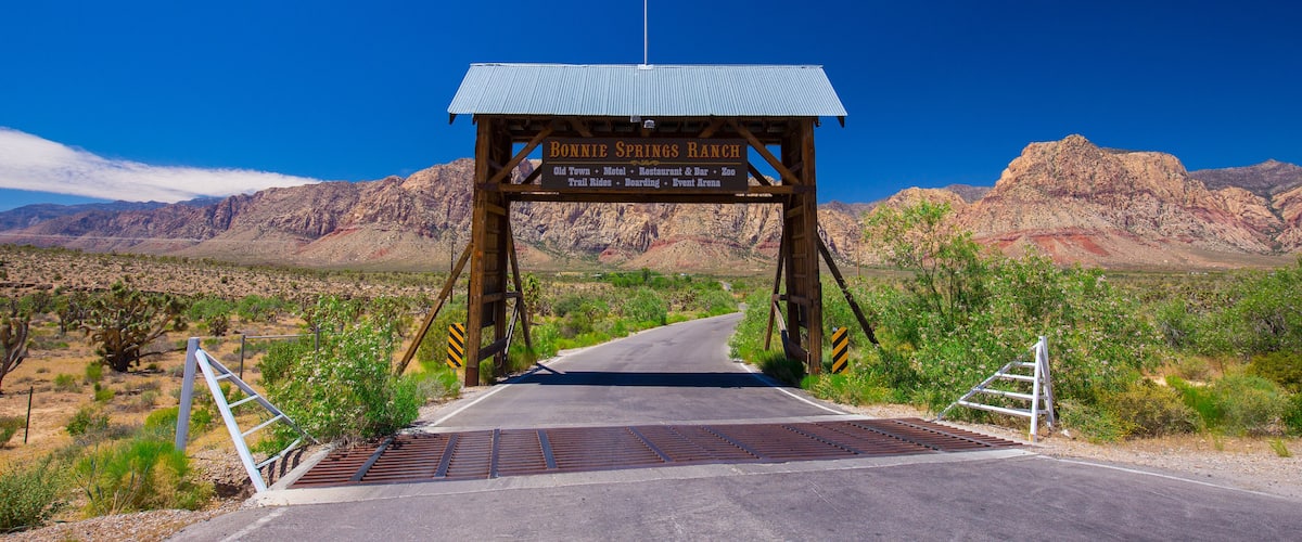 A wooden archway with a sign reading "Bonnie Springs Ranch" and a road leading through a desert landscape with mountains in the background.
