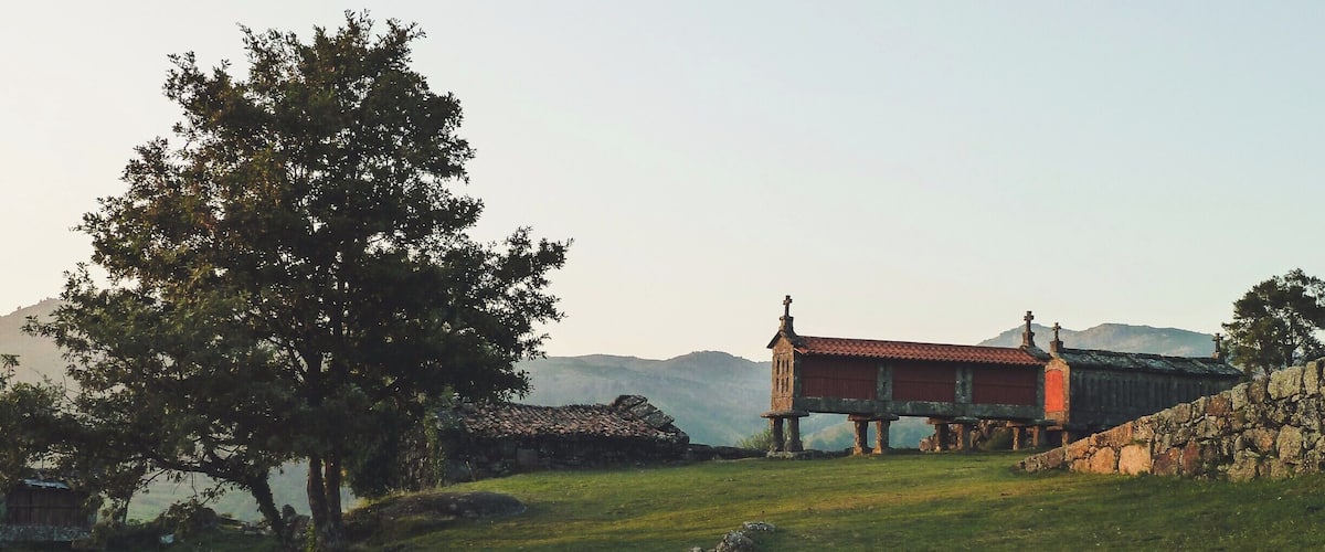 A historic building with a red roof, situated on a grassy hill with a stone wall and a large tree in the foreground.