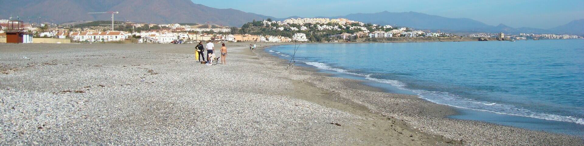 A pebble beach with a clear blue sea, a few people, and buildings in the background.