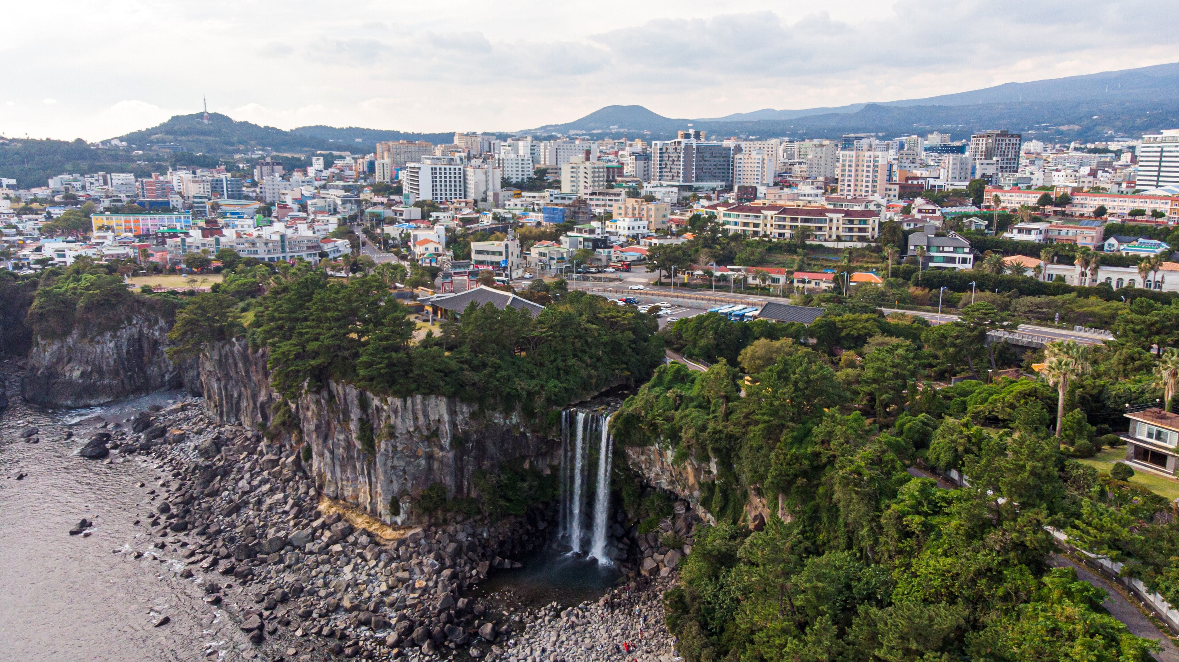 A coastal cityscape with a prominent waterfall, dense greenery, and a mountainous backdrop.