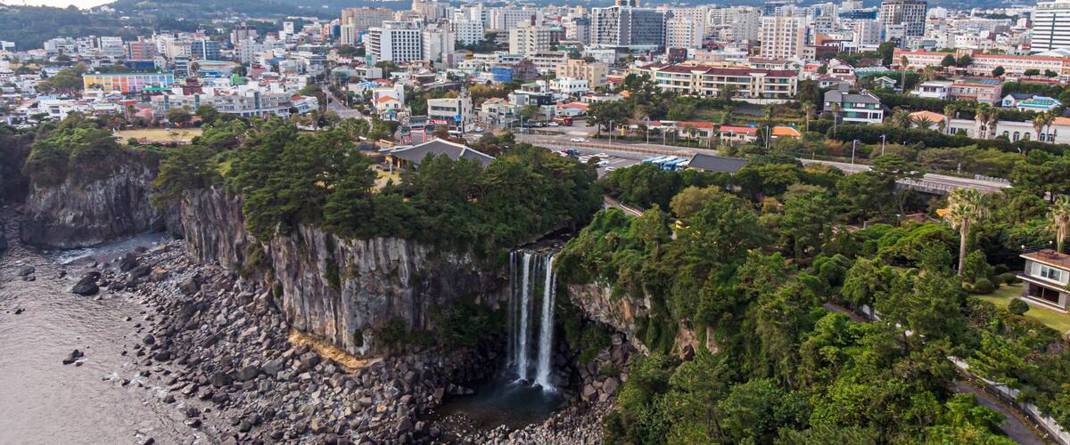 A coastal cityscape with a prominent waterfall, dense greenery, and a mountainous backdrop.
