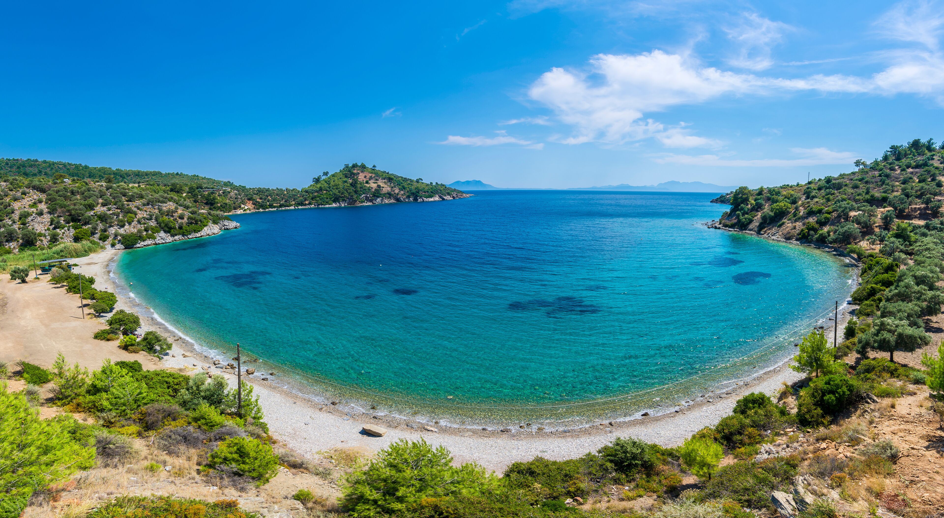 A coastal landscape with a clear blue sea, sandy beach, and surrounding greenery.