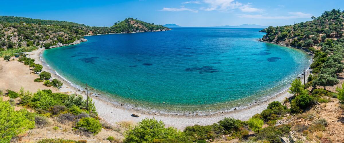 A coastal landscape with a clear blue sea, sandy beach, and surrounding greenery.