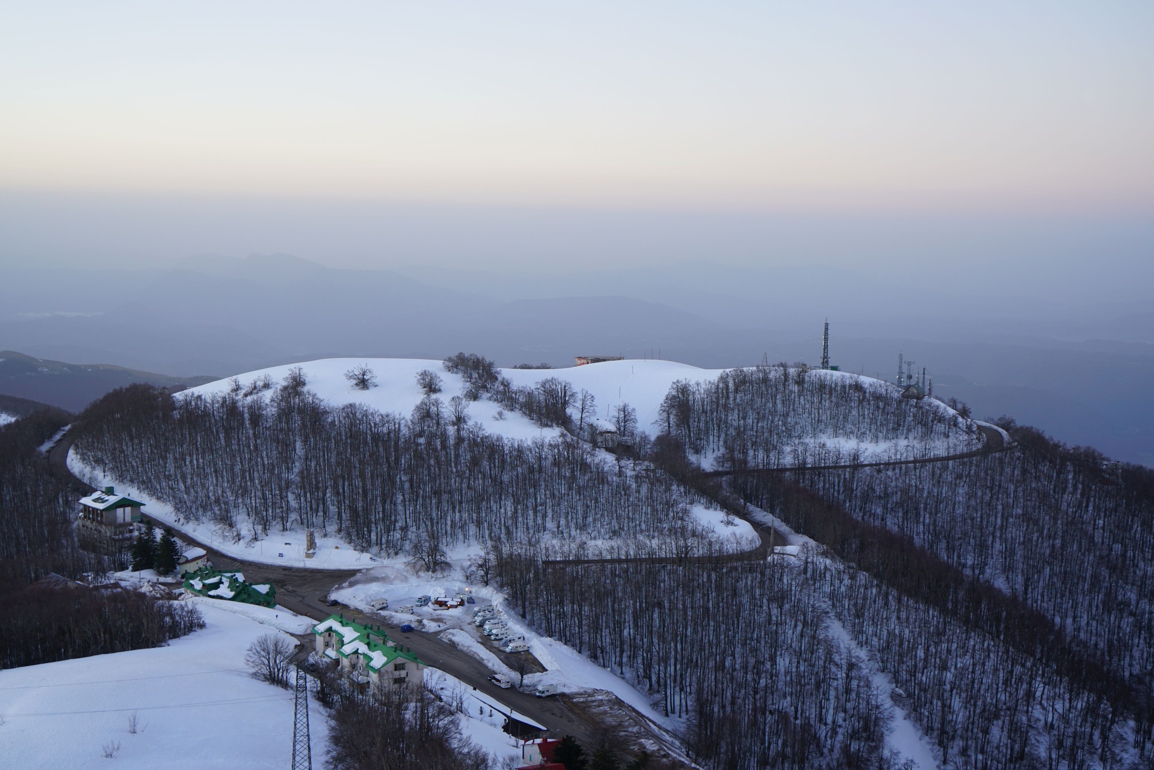 A snow-covered mountainous landscape with a winding road, buildings, and a transmission tower.