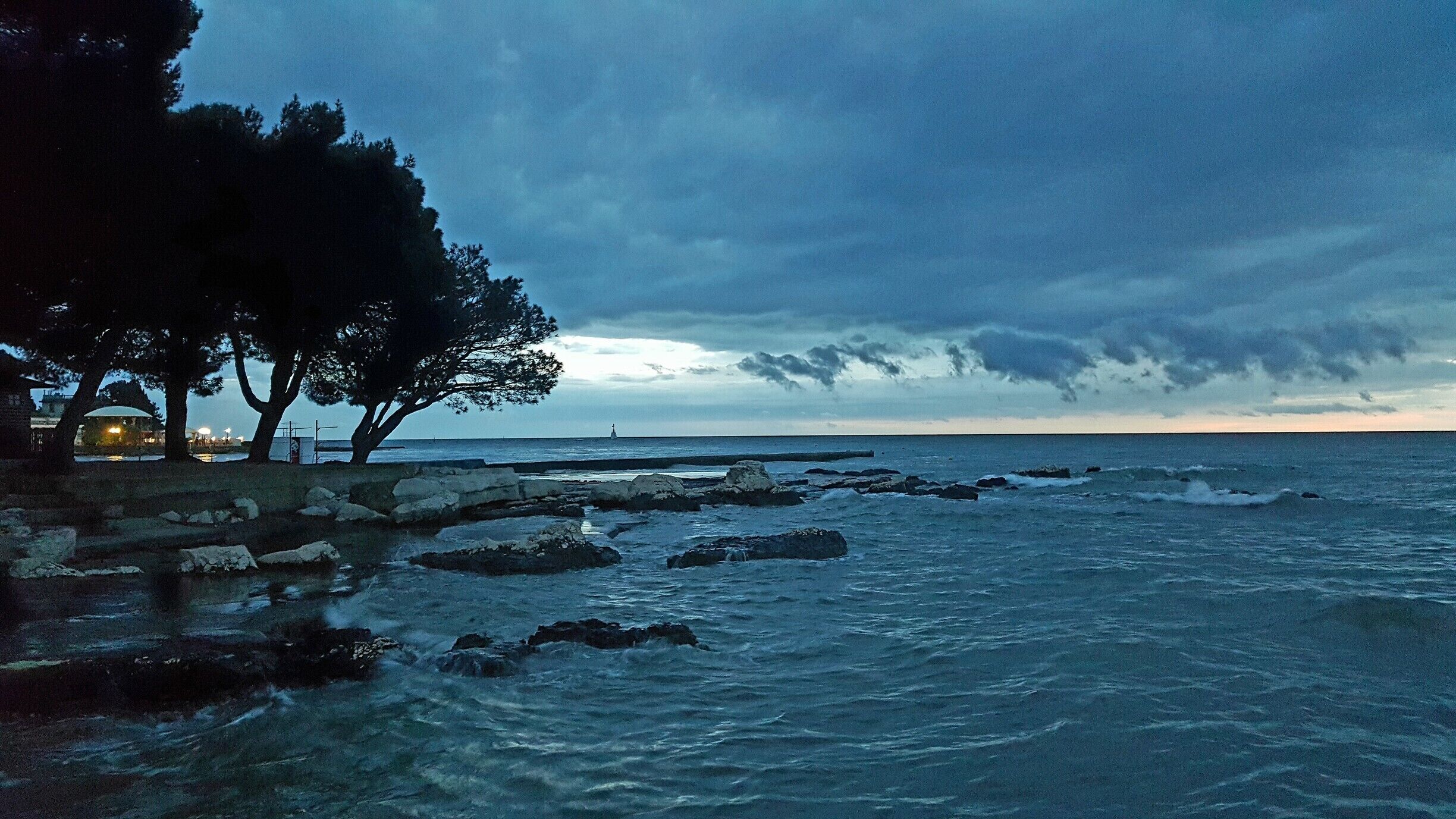 A rocky shoreline with trees and a lighthouse in the distance.