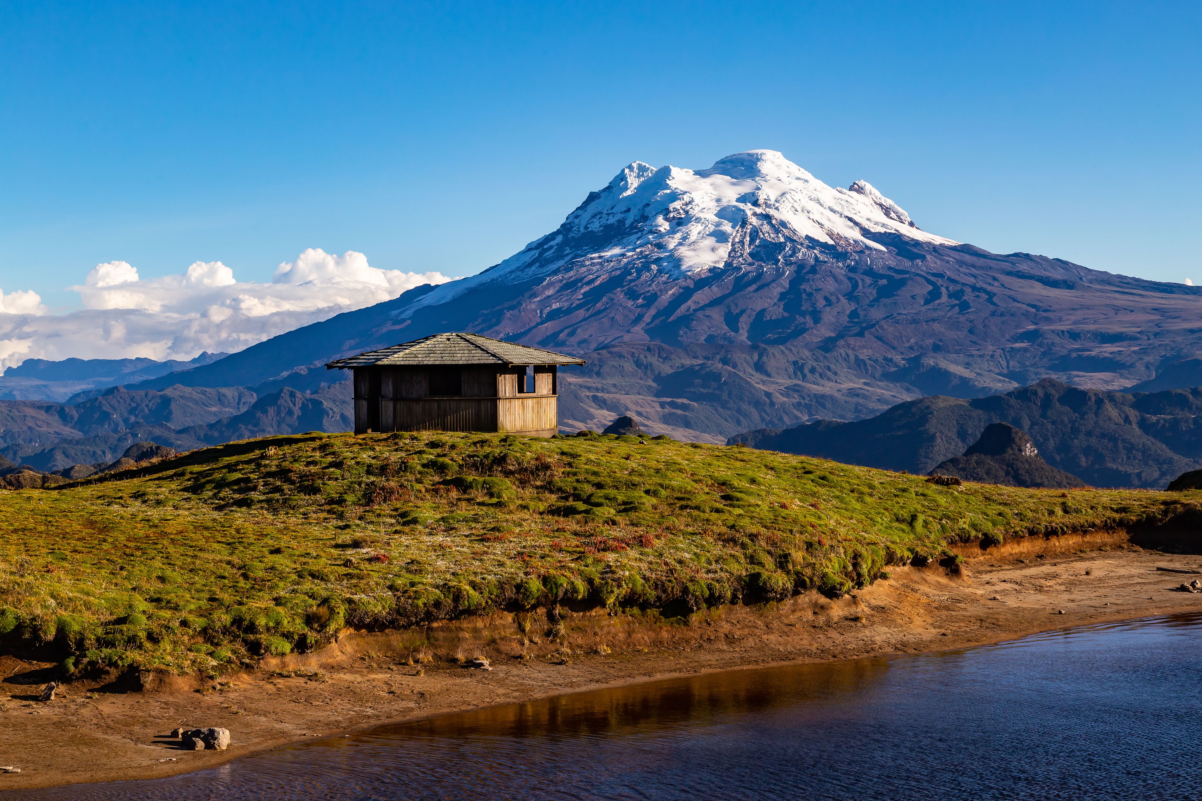 A small wooden hut on a grassy hill with a snow-capped mountain in the background.