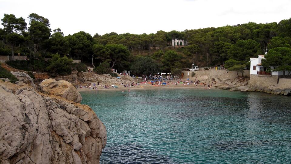A coastal beach with clear turquoise water, rocky cliffs, and a white building in the background.