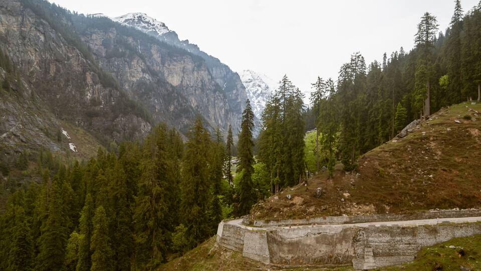 A mountainous landscape with dense forests, a concrete structure, and snow-capped peaks.