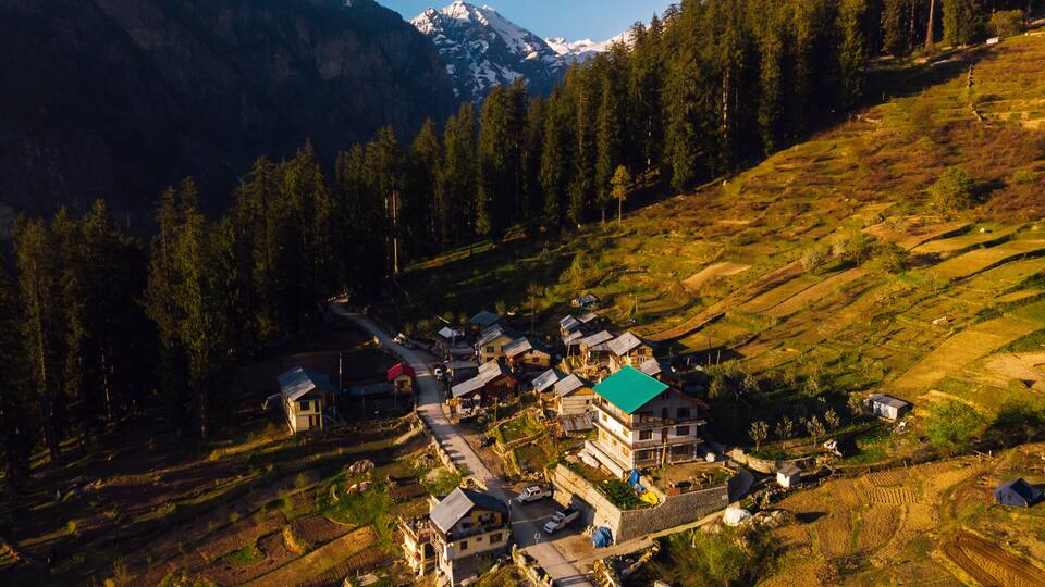 A mountainous village with terraced fields, houses with green roofs, and snow-capped peaks in the background.