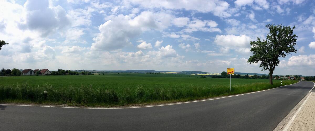 A rural road curving through green fields with houses in the distance and a clear sky.
