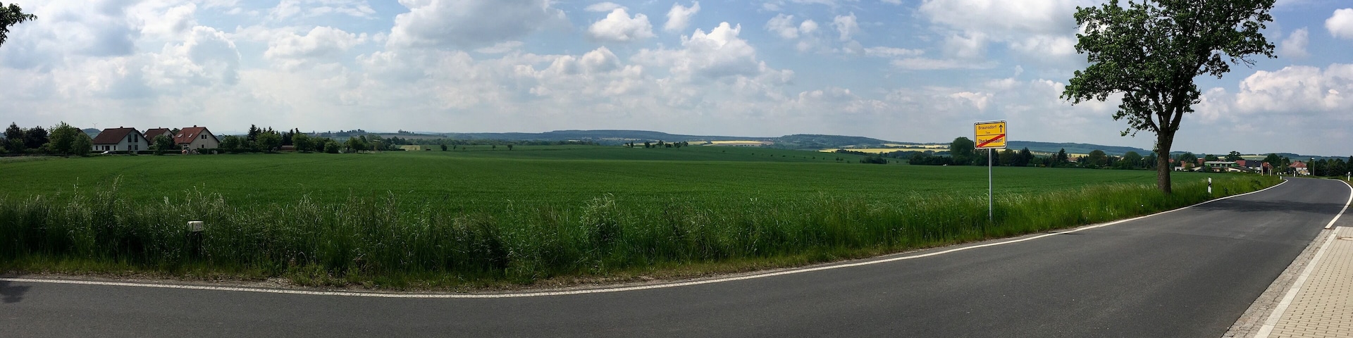 A rural road curving through green fields with houses in the distance and a clear sky.