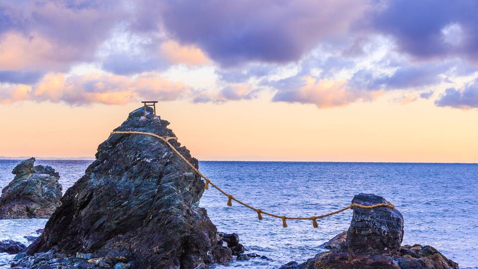 A rocky coastline with a rope bridge over the sea, a wooden structure on top of a rock, and a sunset sky.