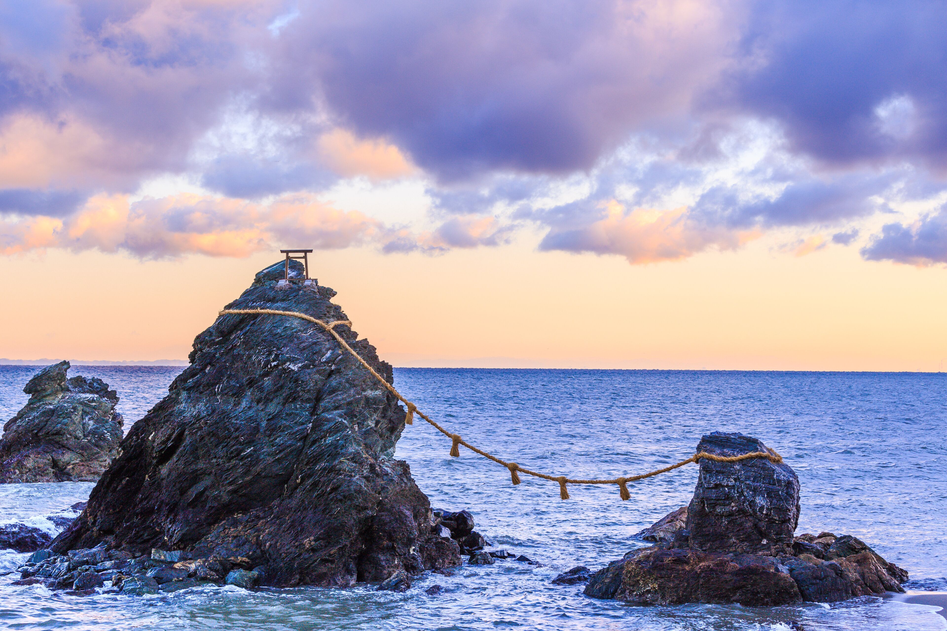 A rocky coastline with a rope bridge over the sea, a wooden structure on top of a rock, and a sunset sky.