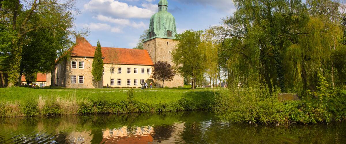 A historic building with a green dome and red roof, surrounded by lush greenery and a calm river.