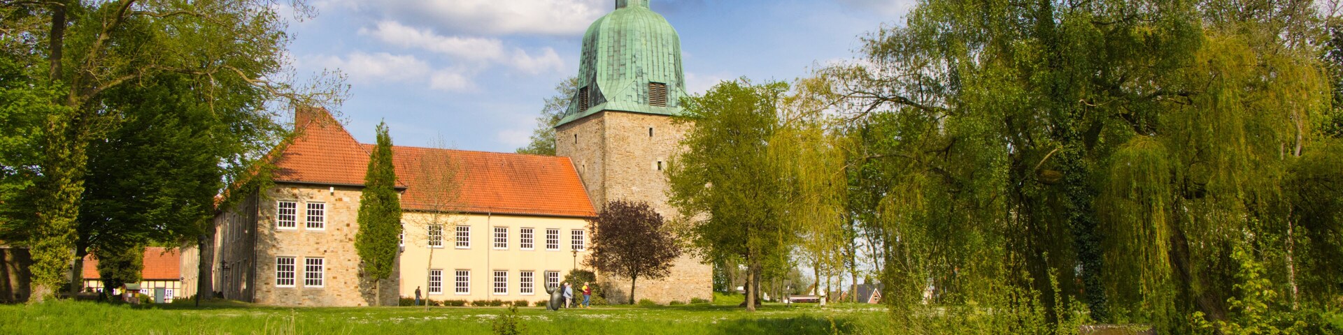 A historic building with a green dome and red roof, surrounded by lush greenery and a calm river.