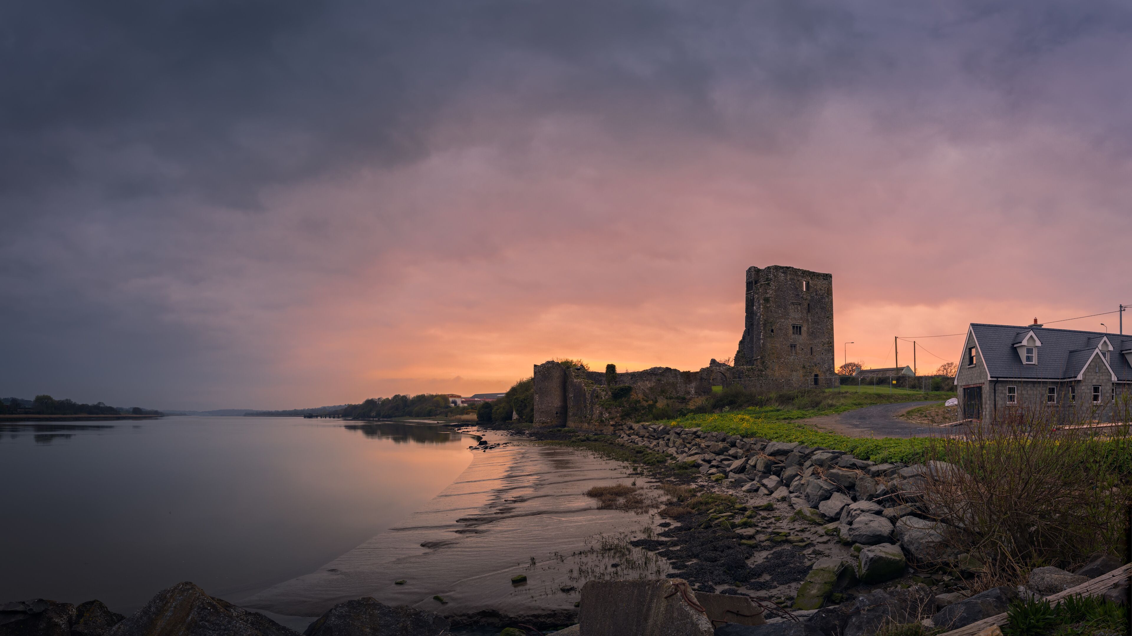 A historic stone tower and a house by a calm river at sunset.