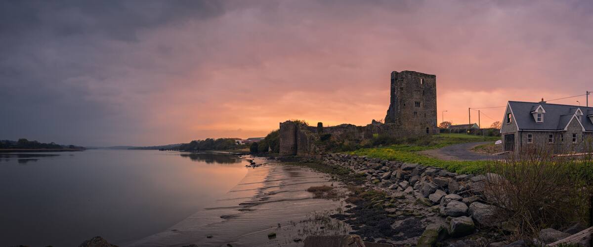 A historic stone tower and a house by a calm river at sunset.