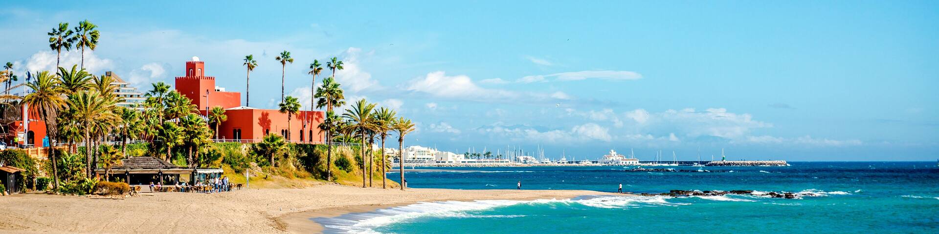 A beach with palm trees, a red building, and a marina with boats.