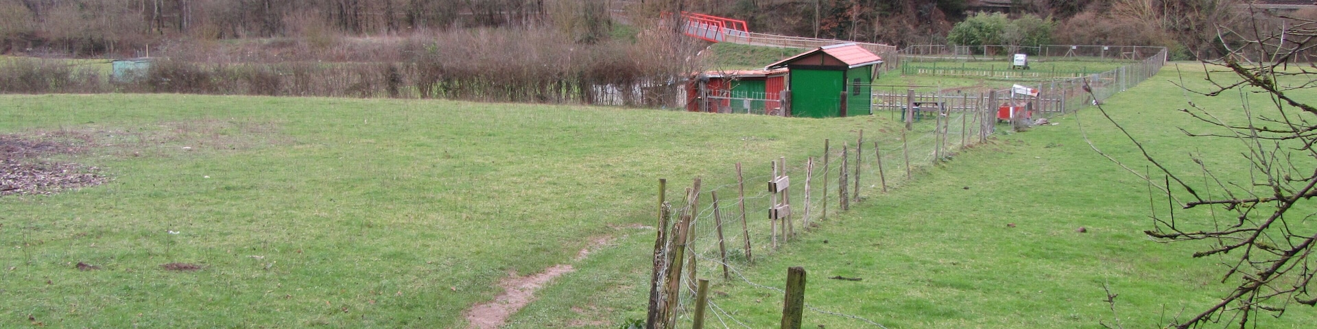 A rural landscape with a green field, a dirt path, a small red and green shed, and a red bridge in the background.