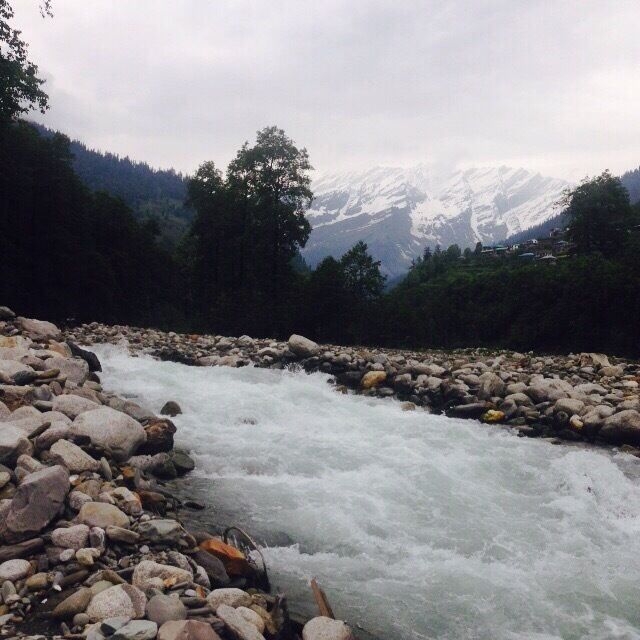 A fast-flowing river with rocks, surrounded by forested hills and snow-capped mountains in the background.