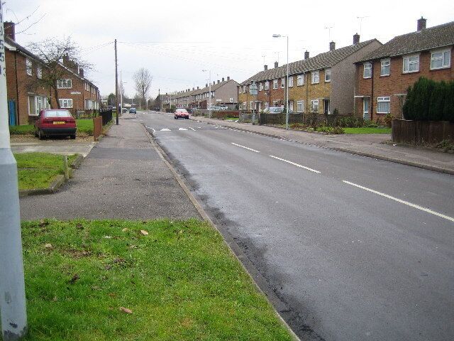 A residential street with houses on both sides, a sidewalk, and cars parked along the road.