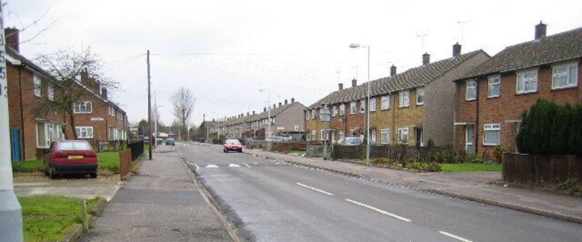 A residential street with houses on both sides, a sidewalk, and cars parked along the road.