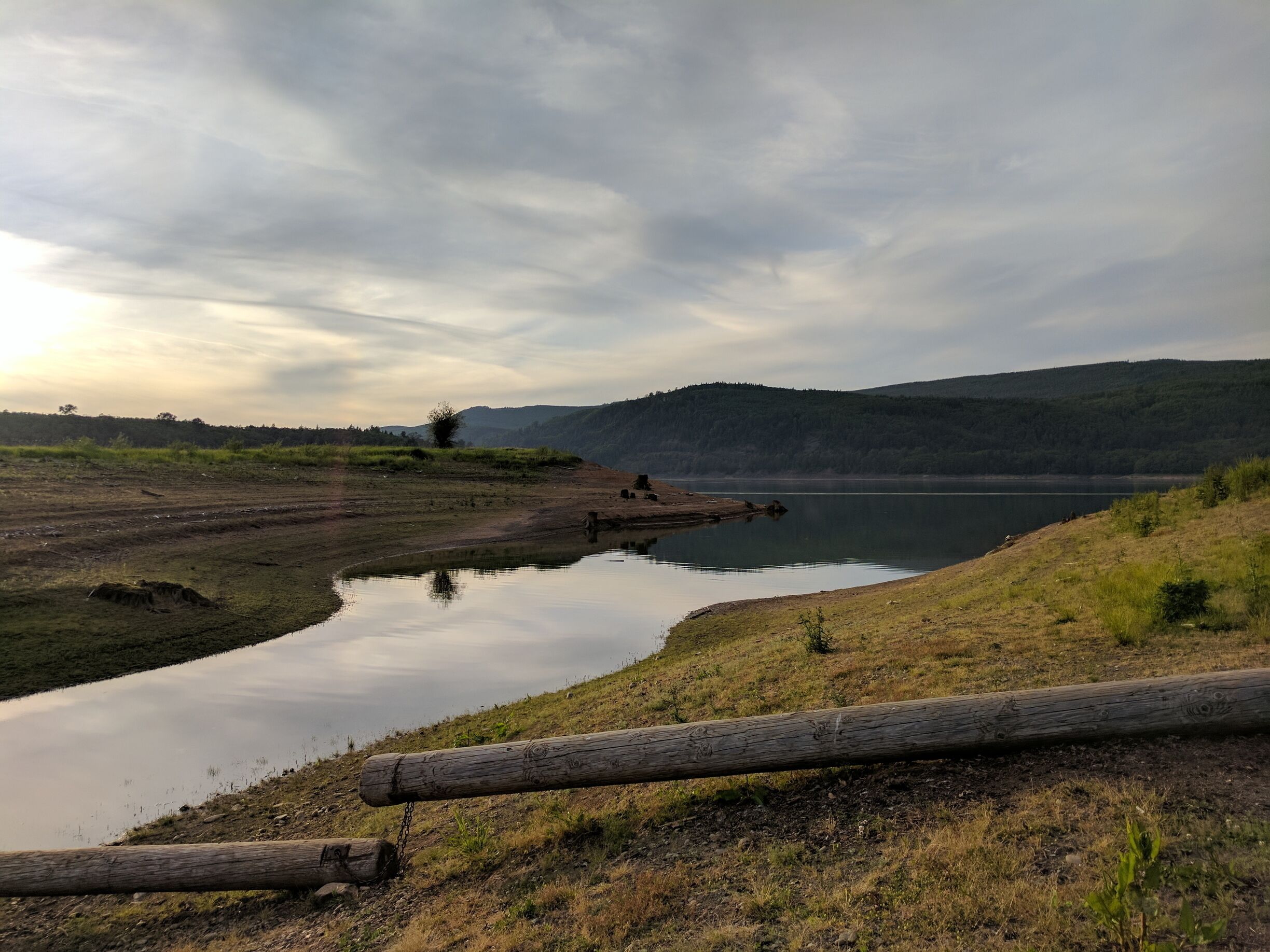 A calm lake with a wooden log in the foreground, surrounded by grassy hills and a cloudy sky.