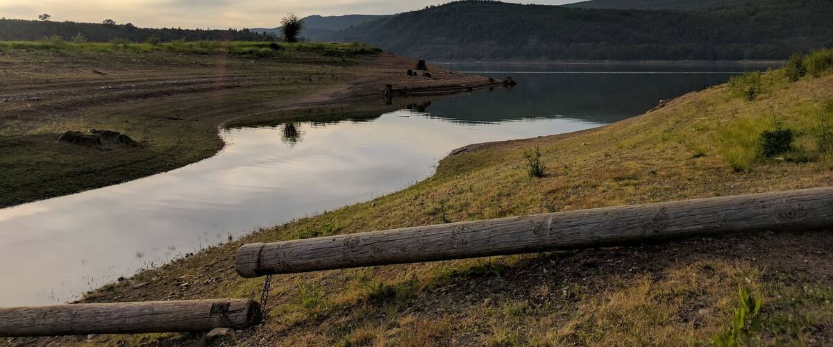 A calm lake with a wooden log in the foreground, surrounded by grassy hills and a cloudy sky.