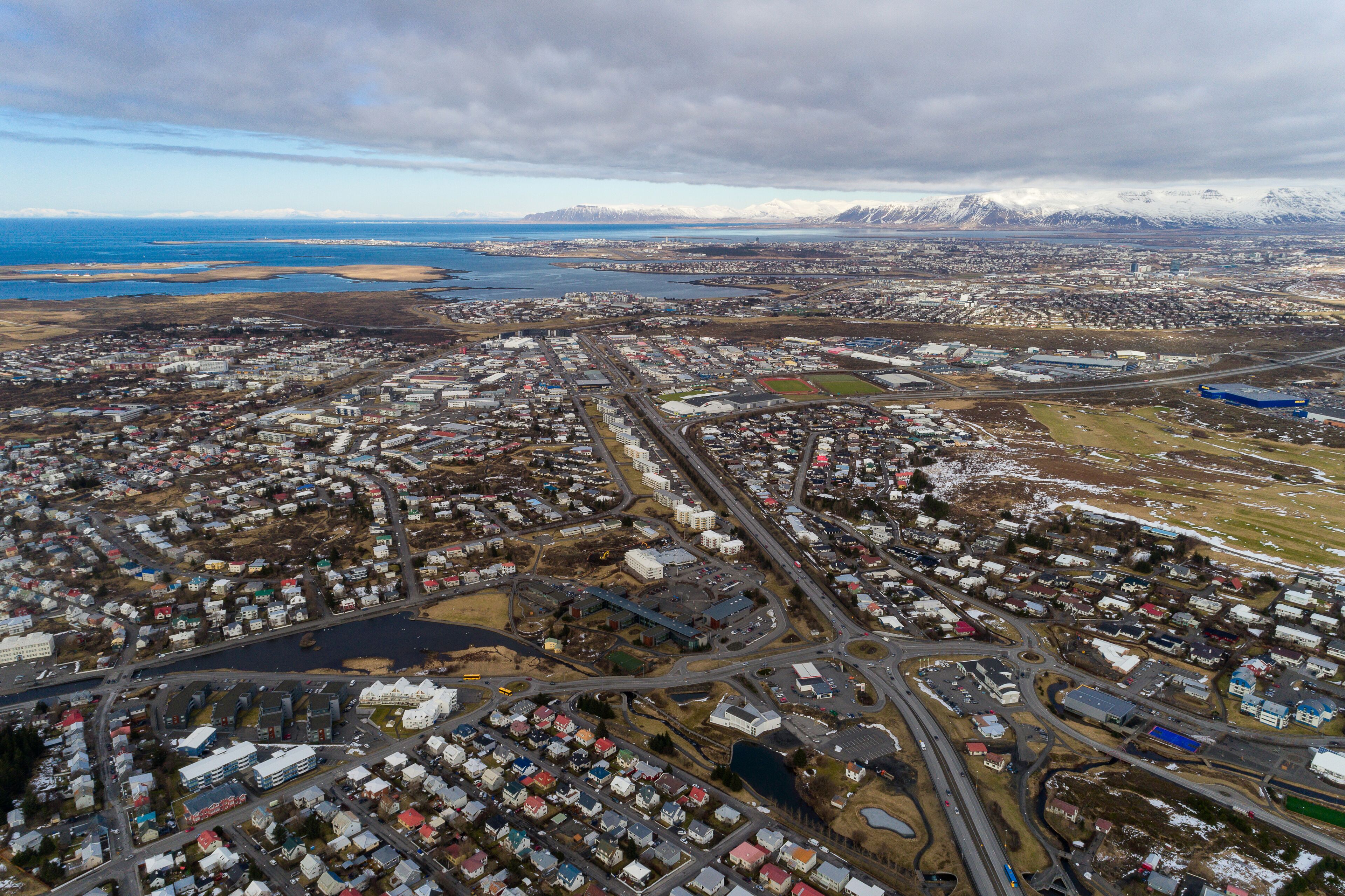 An aerial view of a coastal town with a mix of residential and industrial buildings, a harbor, and snow-capped mountains in the background.