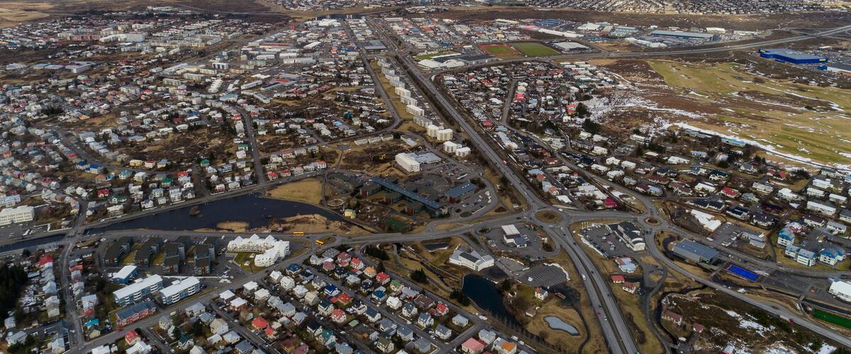 An aerial view of a coastal town with a mix of residential and industrial buildings, a harbor, and snow-capped mountains in the background.