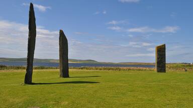 Three standing stones in a grassy field with a lake and hills in the background.