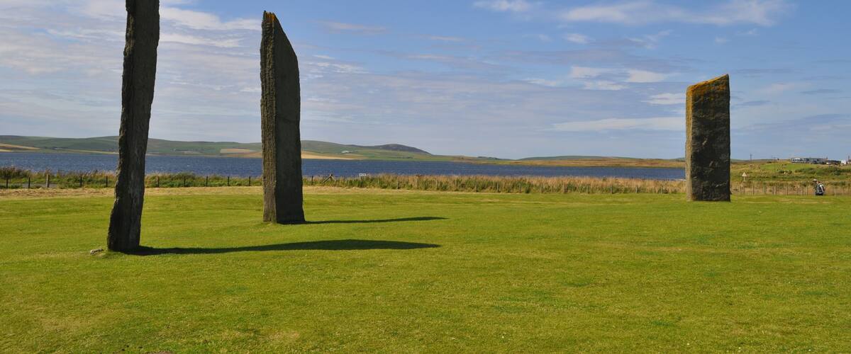 Three standing stones in a grassy field with a lake and hills in the background.
