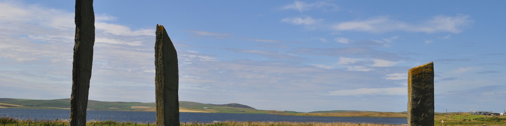 Three standing stones in a grassy field with a lake and hills in the background.