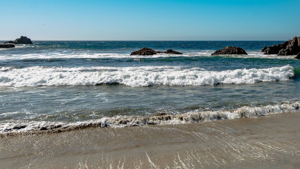A beach with waves crashing onto the shore, rocky outcrops, and a clear blue sky.