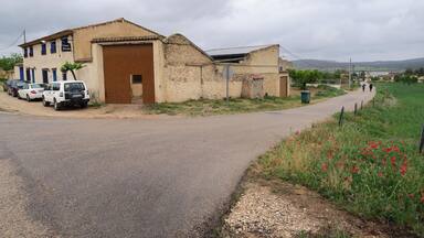 A rural road with parked cars, a building with a large wooden door, and a field of red poppies.