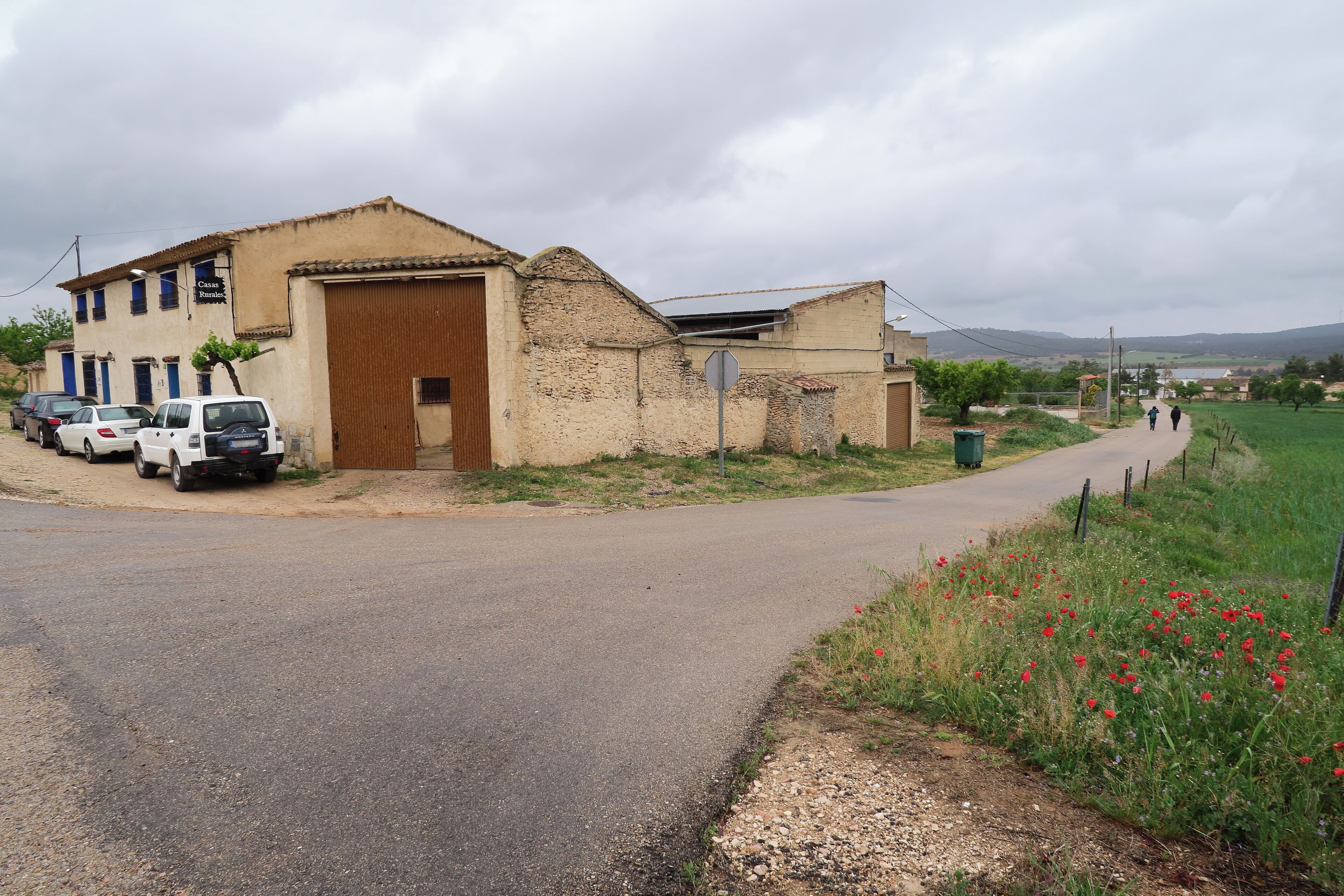 A rural road with parked cars, a building with a large wooden door, and a field of red poppies.