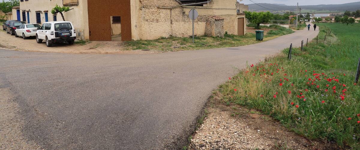 A rural road with parked cars, a building with a large wooden door, and a field of red poppies.