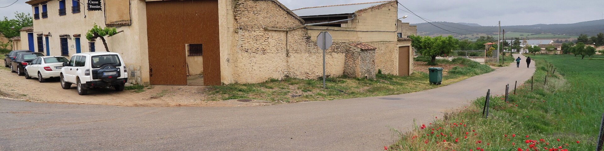 A rural road with parked cars, a building with a large wooden door, and a field of red poppies.