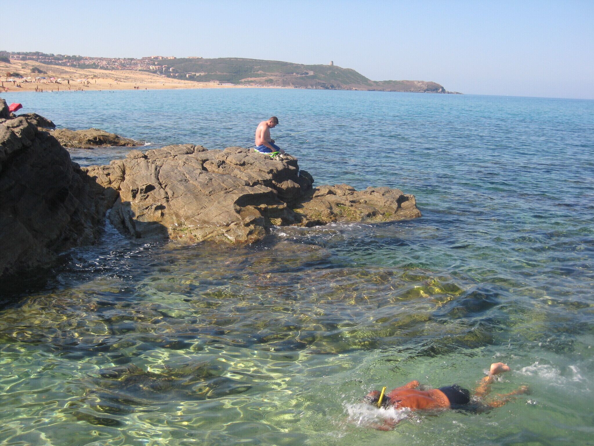 A rocky shoreline with clear water, a swimmer, and a person fishing.