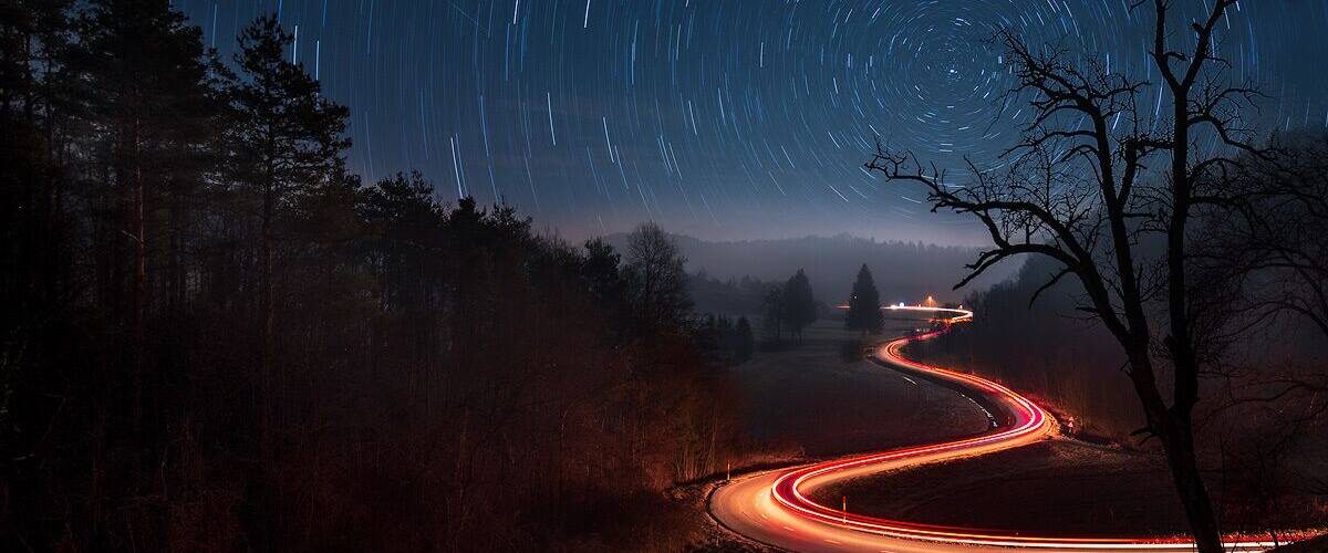 A winding road at night with star trails in the sky.