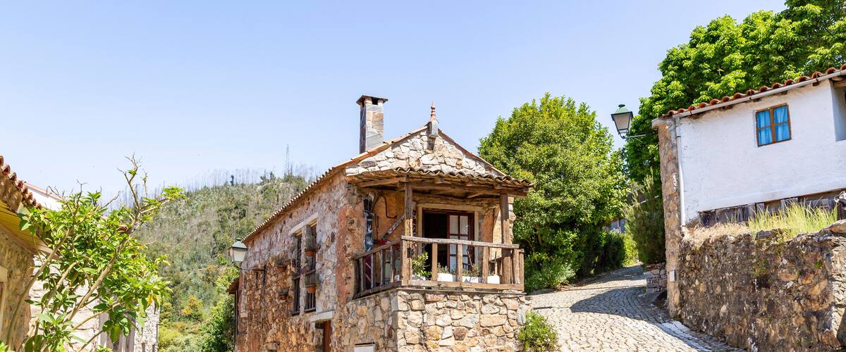 A cobblestone street leading to a stone house with a tiled roof and a wooden balcony.