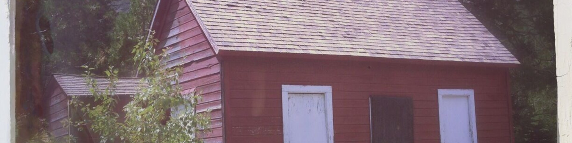 Una cabaña de madera roja con contraventanas blancas, rodeada de árboles y terreno rocoso.