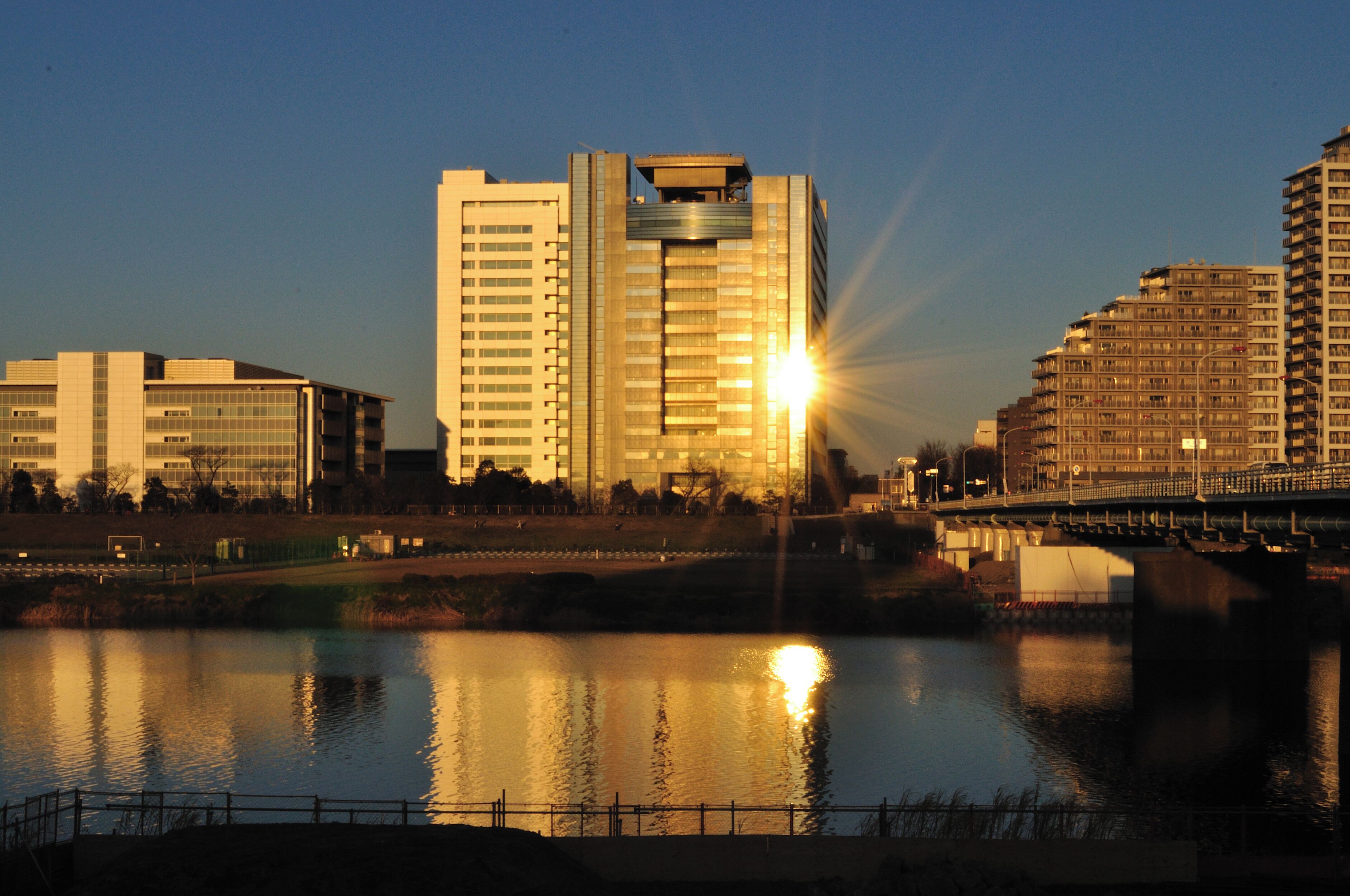 A cityscape with modern buildings and a river reflecting the sunset.