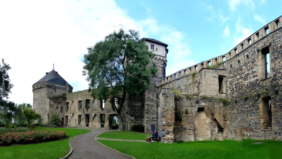 A historic stone castle with a tower and arched windows, surrounded by a well-maintained garden and a paved pathway.